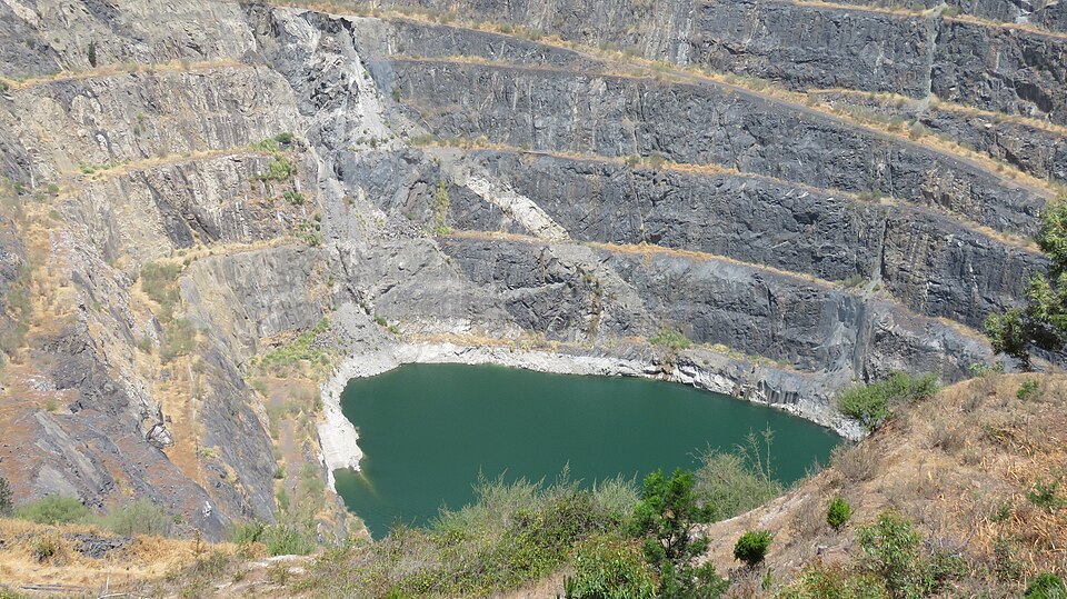 A photo of Greenbushes lithium mine in Australia. It looks like a deep pit of grey rock terraces with a green lake at the bottom. 