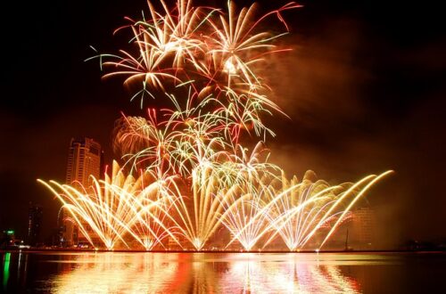 FIreworks in gold, red, and green lighting up the sky over a water source. There are fireworks that look like gold fountains at ground level.