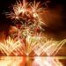 FIreworks in gold, red, and green lighting up the sky over a water source. There are fireworks that look like gold fountains at ground level.
