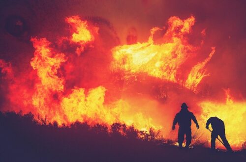 A photo of two firefighters on a hill fighting a wall of huge flames.
