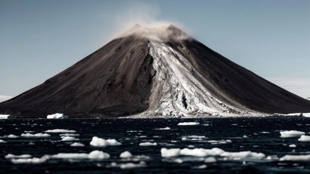 A photo of a volcanic island showing a landslide of rock and ice pouring down the face.