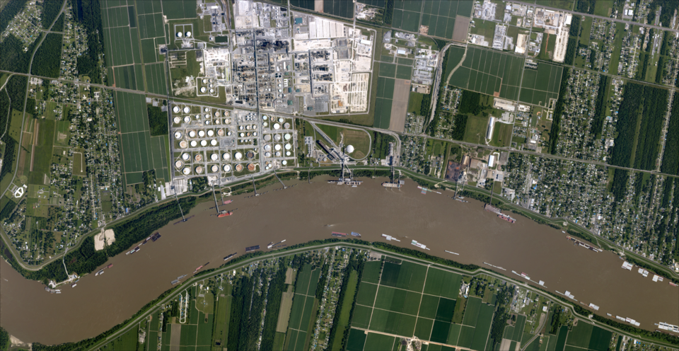 An image of a large oil refinery on the Mississippi River in an area known as cancer alley. The river is dark brown with pollution, and homes are shown nearly. 