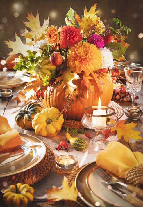 A greener Thanksgiving table decorated with an orange pumpkin filled with colorful flowers, berries, green leaves, and fall leaves. It is surrounded by gourds and leaves.