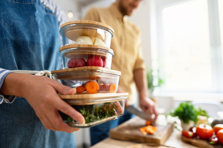 A couple in a kitchen with one person holding 4 glass leftover containers with bamboo lids. The containers are filled with vegetables.
